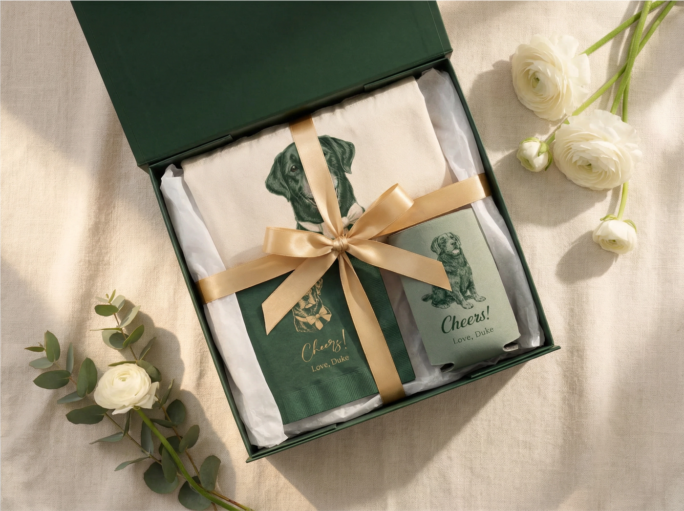 Gift set with a bottle and glasses in a box with a gold ribbon, surrounded by flowers on a light background.