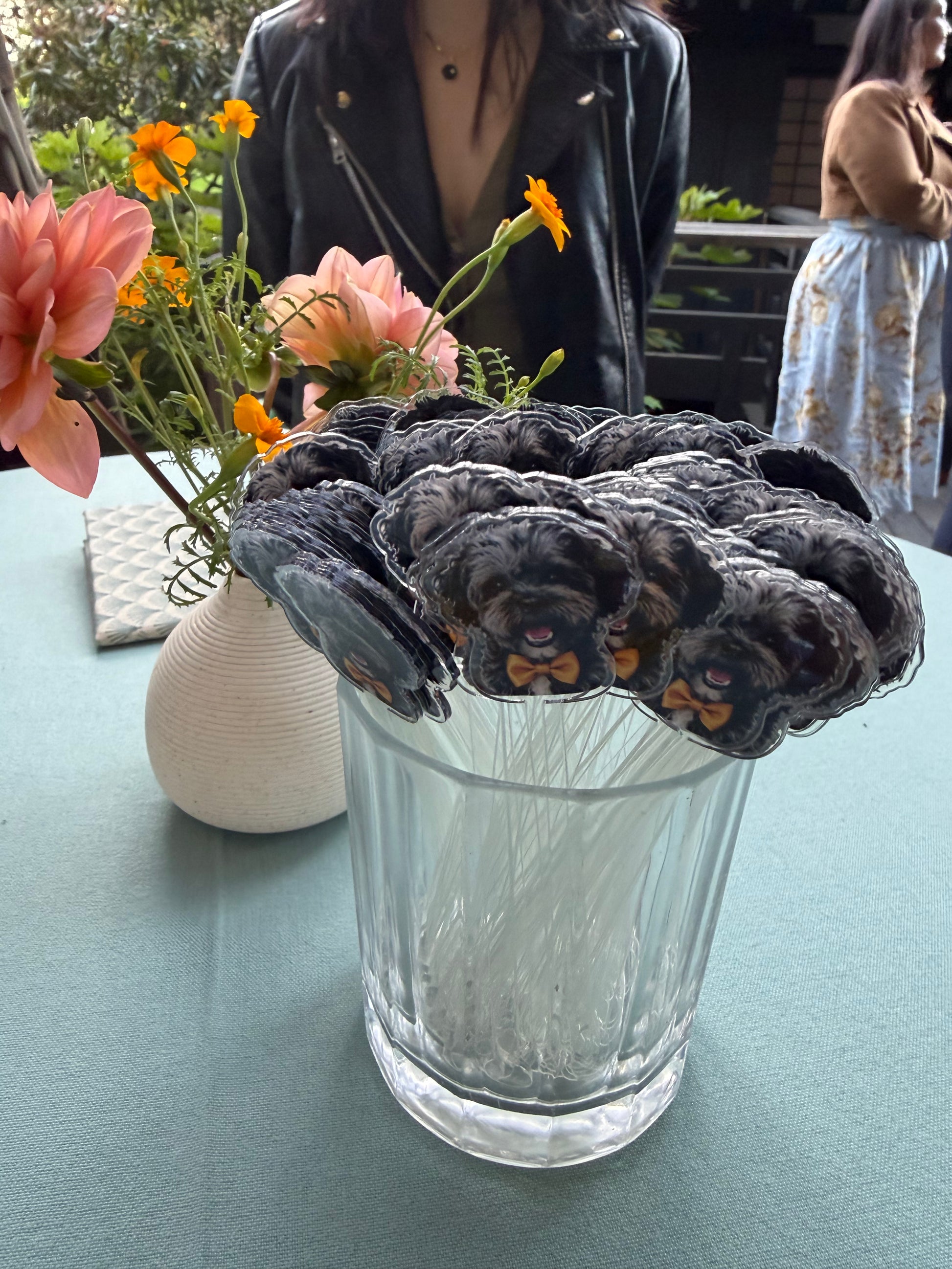 Black dog-shaped cocktail stirrers in a silver cup beside orange flowers on a teal tablecloth