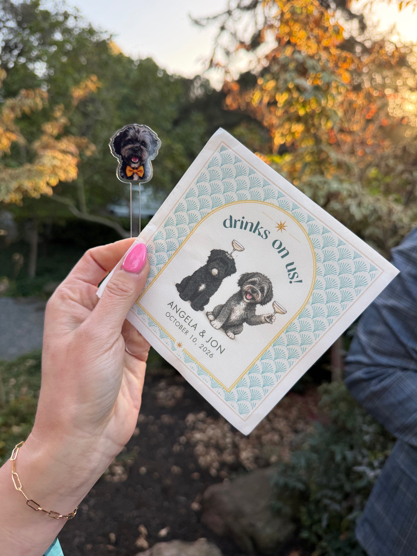 Hand holding a custom Drinks on us napkin with two dog portraits outdoors at a wedding