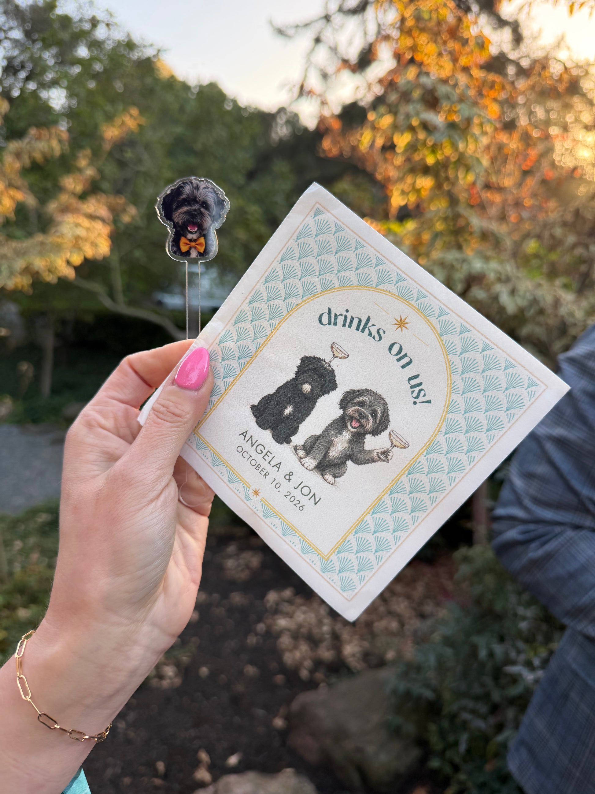 Hand holding a custom Drinks on us napkin with two dog portraits outdoors at a wedding