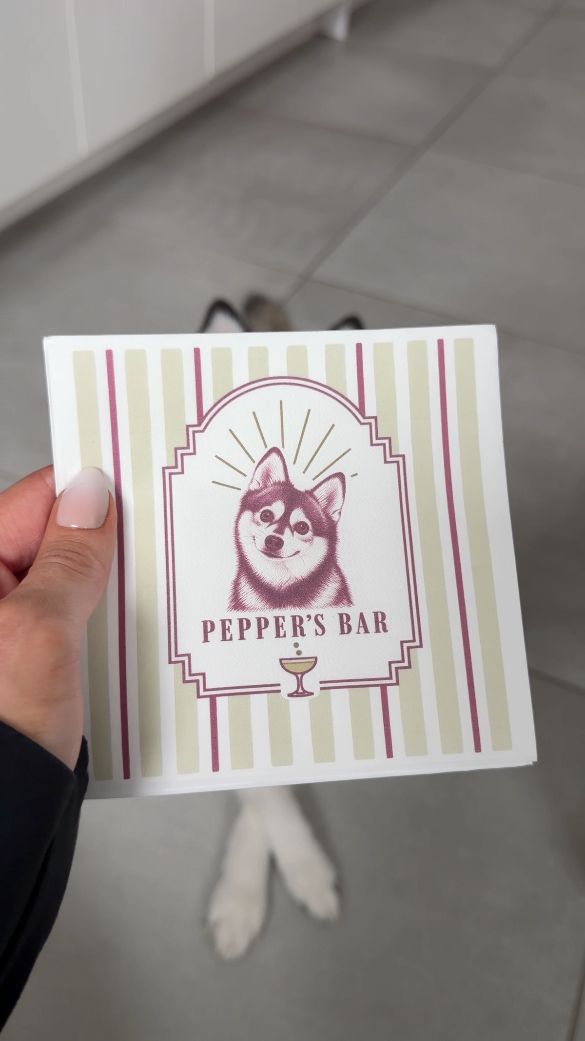 Hand holding a custom cocktail napkin with a husky dog illustration and Pepper's Bar text, with a husky on the floor