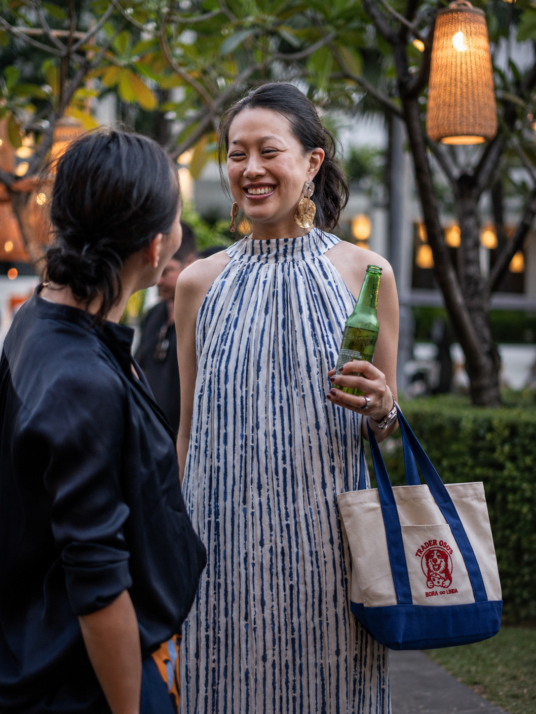 Woman in a blue and white dress holding a bottle, standing outdoors with trees and lights in the background.