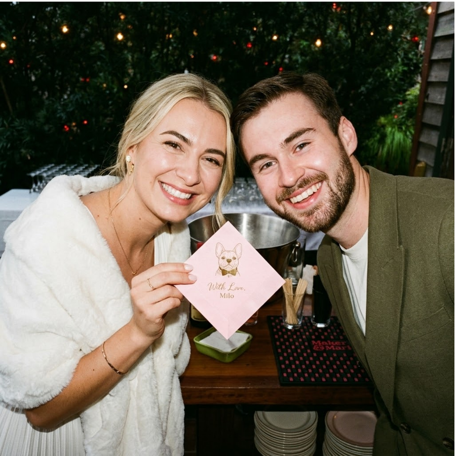 Man and woman holding a pink napkin with a dog's paw print at a festive outdoor event.