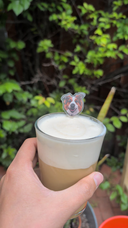 Hand holding a glass of iced coffee with a bear-shaped straw against a green leafy background