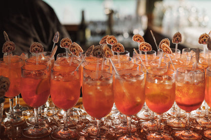 A close-up row of stemmed glasses filled with a bright orange-red cocktail or iced drink, each topped with a decorative spiral garnish on a straw, set against a softly blurred outdoor background.